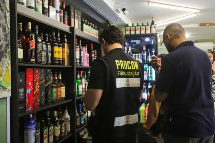 Agente do Procon de Florianópolis realiza fiscalização em bar da capital, observando prateleiras com garrafas de bebidas alcoólicas. A ação visa prevenir a venda de produtos adulterados, especialmente frente aos casos recentes de intoxicação por metanol.