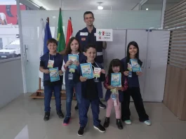 Crianças posam com o supervisor Silvio Dias, da Regional do Imetro de Tubarão, segurando cartilhas do Projeto Fiscal Mirim, destacando a iniciativa de ensino sobre fiscalização e metrologia.