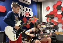 Alunos da School of Rock Floripa se apresentam: um toca baixo, outro guitarra e, ao fundo, aluno na bateria, mostrando a prática musical em grupo.