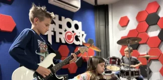 Alunos da School of Rock Floripa se apresentam: um toca baixo, outro guitarra e, ao fundo, aluno na bateria, mostrando a prática musical em grupo.