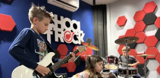 Alunos da School of Rock Floripa se apresentam: um toca baixo, outro guitarra e, ao fundo, aluno na bateria, mostrando a prática musical em grupo.