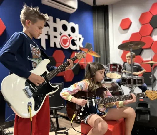 Alunos da School of Rock Floripa se apresentam: um toca baixo, outro guitarra e, ao fundo, aluno na bateria, mostrando a prática musical em grupo.