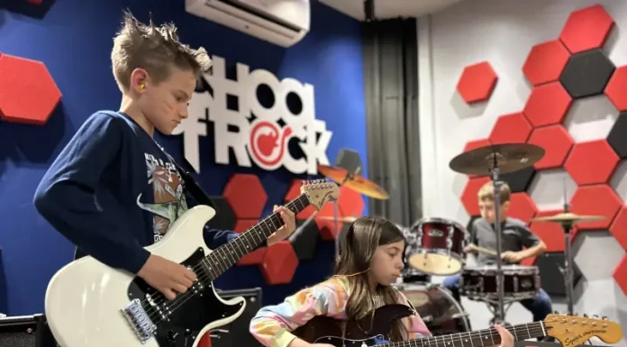 Alunos da School of Rock Floripa se apresentam: um toca baixo, outro guitarra e, ao fundo, aluno na bateria, mostrando a prática musical em grupo.