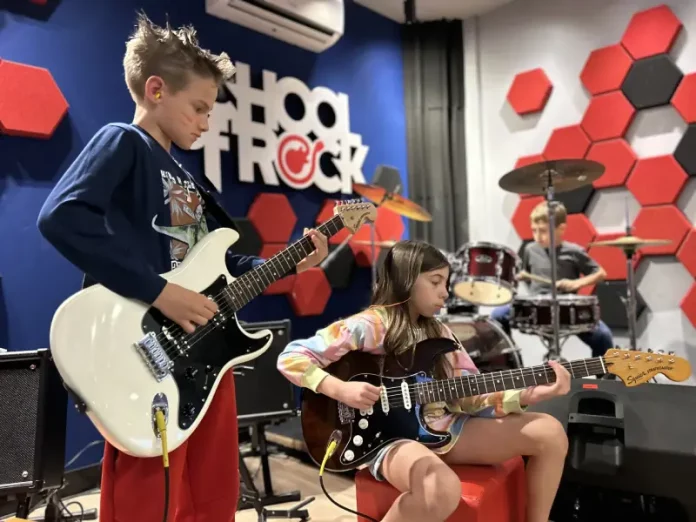 Alunos da School of Rock Floripa se apresentam: um toca baixo, outro guitarra e, ao fundo, aluno na bateria, mostrando a prática musical em grupo.