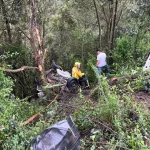 Bombeiro com uniforme amarelo e capacete avalia os destroços de um veículo que caiu em uma ribanceira cercada por mata densa, após grave colisão na BR-282 em Bom Retiro (SC). Ao fundo, um homem observa a cena. Partes do carro estão espalhadas entre a vegetação, indicando a força do impacto.