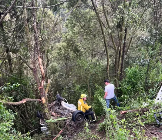 Bombeiro com uniforme amarelo e capacete avalia os destroços de um veículo que caiu em uma ribanceira cercada por mata densa, após grave colisão na BR-282 em Bom Retiro (SC). Ao fundo, um homem observa a cena. Partes do carro estão espalhadas entre a vegetação, indicando a força do impacto.