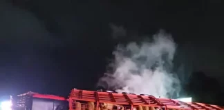Acidente com caminhão carregado de botijões de gás tombado no vão central da pista em chamas, com outro caminhão colidido na traseira do veículo tombado.