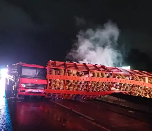 Acidente com caminhão carregado de botijões de gás tombado no vão central da pista em chamas, com outro caminhão colidido na traseira do veículo tombado.