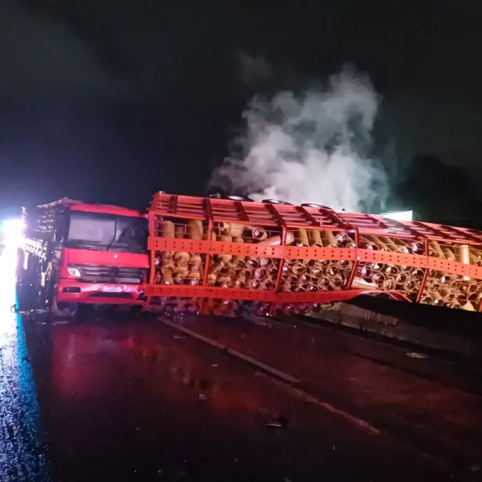 Acidente com caminhão carregado de botijões de gás tombado no vão central da pista em chamas, com outro caminhão colidido na traseira do veículo tombado.
