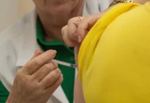 Profissional de saúde aplica vacina no braço de uma pessoa vestida com camiseta amarela. A imagem ilustra a campanha de multivacinação, que busca atualizar a caderneta vacinal de crianças e adolescentes.