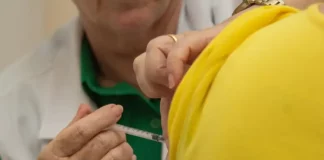 Profissional de saúde aplica vacina no braço de uma pessoa vestida com camiseta amarela. A imagem ilustra a campanha de multivacinação, que busca atualizar a caderneta vacinal de crianças e adolescentes.