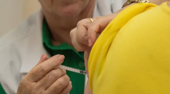 Profissional de saúde aplica vacina no braço de uma pessoa vestida com camiseta amarela. A imagem ilustra a campanha de multivacinação, que busca atualizar a caderneta vacinal de crianças e adolescentes.