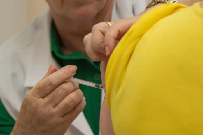 Profissional de saúde aplica vacina no braço de uma pessoa vestida com camiseta amarela. A imagem ilustra a campanha de multivacinação, que busca atualizar a caderneta vacinal de crianças e adolescentes.