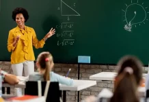 Professora em sala de aula diante do quadro com questões de matemática, olhando para os alunos. A imagem ilustra o processo seletivo para professores da rede municipal de São José.