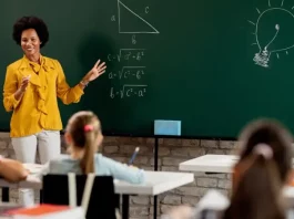 Professora em sala de aula diante do quadro com questões de matemática, olhando para os alunos. A imagem ilustra o processo seletivo para professores da rede municipal de São José.