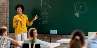 Professora em sala de aula diante do quadro com questões de matemática, olhando para os alunos. A imagem ilustra o processo seletivo para professores da rede municipal de São José.