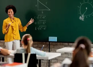 Professora em sala de aula diante do quadro com questões de matemática, olhando para os alunos. A imagem ilustra o processo seletivo para professores da rede municipal de São José.