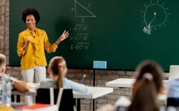 Professora em sala de aula diante do quadro com questões de matemática, olhando para os alunos. A imagem ilustra o processo seletivo para professores da rede municipal de São José.