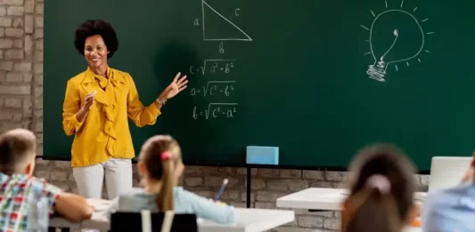 Professora em sala de aula diante do quadro com questões de matemática, olhando para os alunos. A imagem ilustra o processo seletivo para professores da rede municipal de São José.