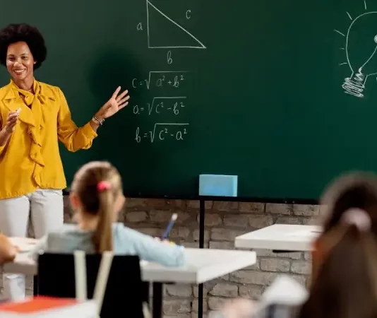 Professora em sala de aula diante do quadro com questões de matemática, olhando para os alunos. A imagem ilustra o processo seletivo para professores da rede municipal de São José.