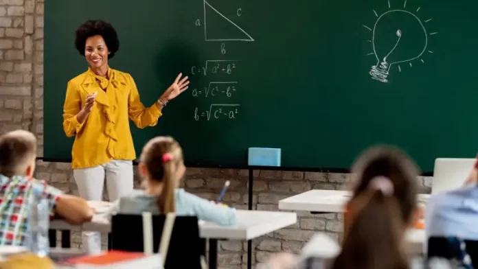 Professora em sala de aula diante do quadro com questões de matemática, olhando para os alunos. A imagem ilustra o processo seletivo para professores da rede municipal de São José.
