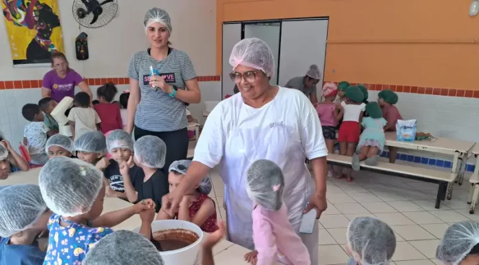 Cozinheira Stela na cozinha do NEIM Mateus de Barros, auxiliando crianças no preparo da massa de bolo, em atividade educativa e prática de alimentação saudável.