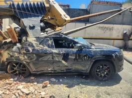 Imagem registrada para documentar os danos causados por um deslizamento de terra ocorrido na manhã de terça-feira, 14 de outubro, no Centro de Florianópolis. A foto mostra uma escavadeira tombada sobre um SUV preto estacionado, com danos significativos na estrutura do veículo. A máquina teria provocado o deslizamento ao operar em um terreno instável. A imagem tem como finalidade ilustrar os efeitos do acidente, demonstrando a gravidade da ocorrência e subsidiar ações de avaliação de risco, segurança e resposta das autoridades envolvidas, como o Corpo de Bombeiros e a Defesa Civil.
