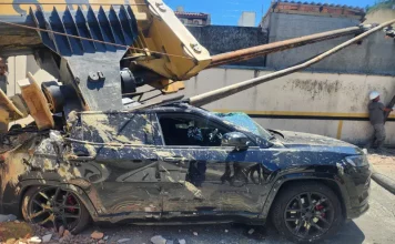 Imagem registrada para documentar os danos causados por um deslizamento de terra ocorrido na manhã de terça-feira, 14 de outubro, no Centro de Florianópolis. A foto mostra uma escavadeira tombada sobre um SUV preto estacionado, com danos significativos na estrutura do veículo. A máquina teria provocado o deslizamento ao operar em um terreno instável. A imagem tem como finalidade ilustrar os efeitos do acidente, demonstrando a gravidade da ocorrência e subsidiar ações de avaliação de risco, segurança e resposta das autoridades envolvidas, como o Corpo de Bombeiros e a Defesa Civil.