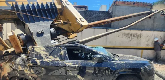 Imagem registrada para documentar os danos causados por um deslizamento de terra ocorrido na manhã de terça-feira, 14 de outubro, no Centro de Florianópolis. A foto mostra uma escavadeira tombada sobre um SUV preto estacionado, com danos significativos na estrutura do veículo. A máquina teria provocado o deslizamento ao operar em um terreno instável. A imagem tem como finalidade ilustrar os efeitos do acidente, demonstrando a gravidade da ocorrência e subsidiar ações de avaliação de risco, segurança e resposta das autoridades envolvidas, como o Corpo de Bombeiros e a Defesa Civil.