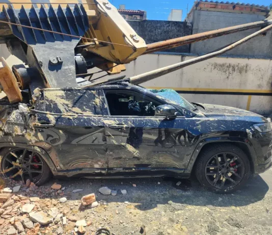 Imagem registrada para documentar os danos causados por um deslizamento de terra ocorrido na manhã de terça-feira, 14 de outubro, no Centro de Florianópolis. A foto mostra uma escavadeira tombada sobre um SUV preto estacionado, com danos significativos na estrutura do veículo. A máquina teria provocado o deslizamento ao operar em um terreno instável. A imagem tem como finalidade ilustrar os efeitos do acidente, demonstrando a gravidade da ocorrência e subsidiar ações de avaliação de risco, segurança e resposta das autoridades envolvidas, como o Corpo de Bombeiros e a Defesa Civil.