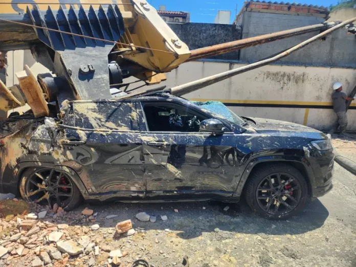 Imagem registrada para documentar os danos causados por um deslizamento de terra ocorrido na manhã de terça-feira, 14 de outubro, no Centro de Florianópolis. A foto mostra uma escavadeira tombada sobre um SUV preto estacionado, com danos significativos na estrutura do veículo. A máquina teria provocado o deslizamento ao operar em um terreno instável. A imagem tem como finalidade ilustrar os efeitos do acidente, demonstrando a gravidade da ocorrência e subsidiar ações de avaliação de risco, segurança e resposta das autoridades envolvidas, como o Corpo de Bombeiros e a Defesa Civil.