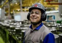 Operário sorrindo em linha de produção, usando capacete, óculos e uniforme, simbolizando o baixo desemprego em Santa Catarina, o menor do país segundo o Censo 2022.