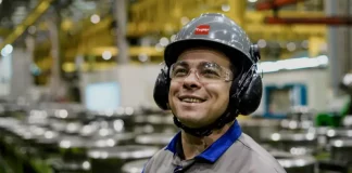 Operário sorrindo em linha de produção, usando capacete, óculos e uniforme, simbolizando o baixo desemprego em Santa Catarina, o menor do país segundo o Censo 2022.