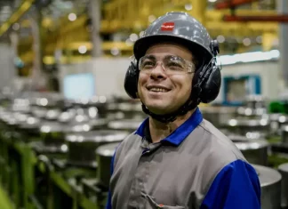 Operário sorrindo em linha de produção, usando capacete, óculos e uniforme, simbolizando o baixo desemprego em Santa Catarina, o menor do país segundo o Censo 2022.