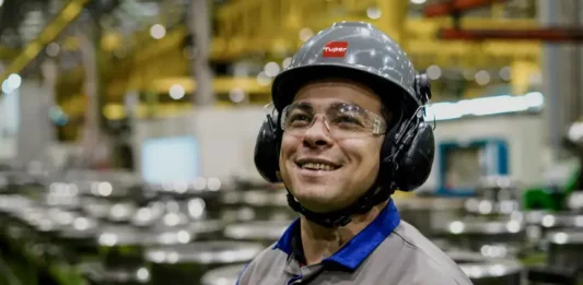 Operário sorrindo em linha de produção, usando capacete, óculos e uniforme, simbolizando o baixo desemprego em Santa Catarina, o menor do país segundo o Censo 2022.