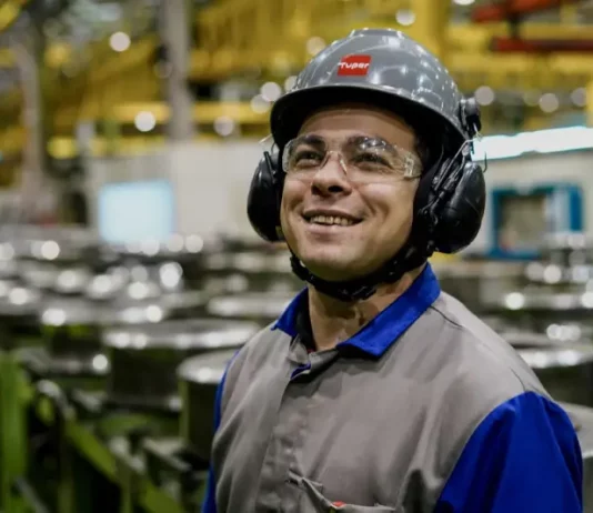Operário sorrindo em linha de produção, usando capacete, óculos e uniforme, simbolizando o baixo desemprego em Santa Catarina, o menor do país segundo o Censo 2022.