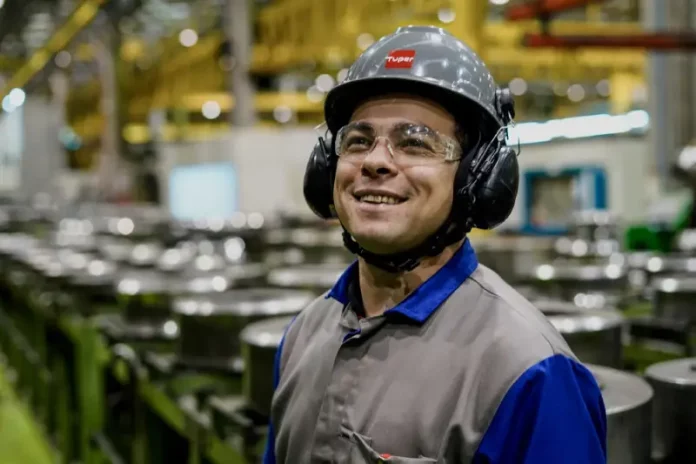 Operário sorrindo em linha de produção, usando capacete, óculos e uniforme, simbolizando o baixo desemprego em Santa Catarina, o menor do país segundo o Censo 2022.