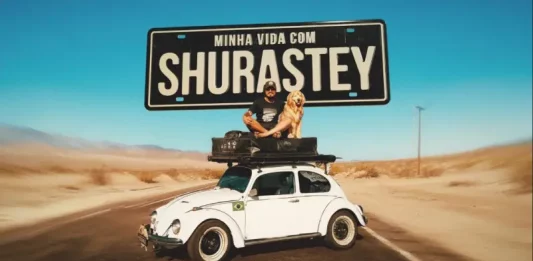 Cena promocional do filme “Minha Vida com Shurastey”, mostrando um homem e seu cachorro sobre um fusca branco em uma estrada desértica, sob uma placa com o título do longa. A imagem é usada para divulgar a produção inspirada na história real de Jesse Kozechen e seu cão Shurastey.