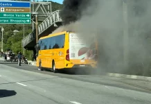 A imagem mostra um ônibus amarelo em chamas após a colisão com uma motocicleta. O registro evidencia a gravidade do acidente, que resultou na morte do motociclista, e a atuação das equipes de resgate diante do incêndio.