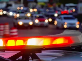 Luzes vermelhas e amarelas de uma viatura policial acesas em primeiro plano, com o trânsito urbano desfocado ao fundo, simbolizando a atuação das forças de segurança no combate aos homicídios e na redução da violência em Santa Catarina.