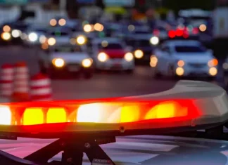 Luzes vermelhas e amarelas de uma viatura policial acesas em primeiro plano, com o trânsito urbano desfocado ao fundo, simbolizando a atuação das forças de segurança no combate aos homicídios e na redução da violência em Santa Catarina.
