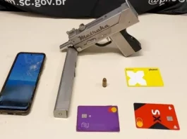 A imagem mostra uma submetralhadora artesanal, um celular e cartões de banco apreendidos durante a operação policial, ilustrando os itens recolhidos pelos agentes.