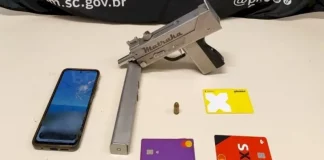 A imagem mostra uma submetralhadora artesanal, um celular e cartões de banco apreendidos durante a operação policial, ilustrando os itens recolhidos pelos agentes.