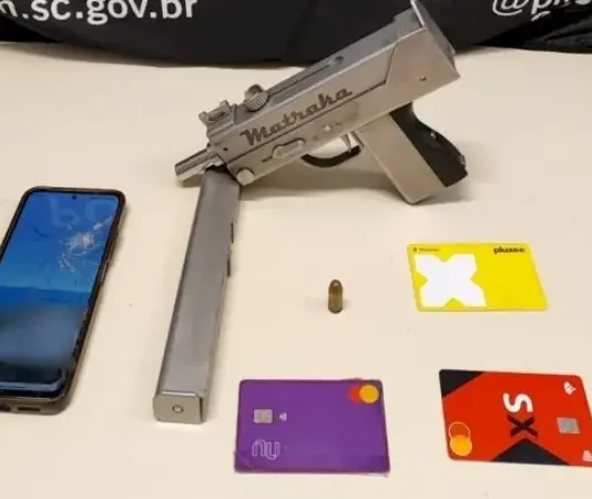 Submetralhadora artesanal é apreendida durante ação policial em São José A imagem mostra uma submetralhadora artesanal, um celular e cartões de banco apreendidos durante a operação policial, ilustrando os itens recolhidos pelos agentes.
