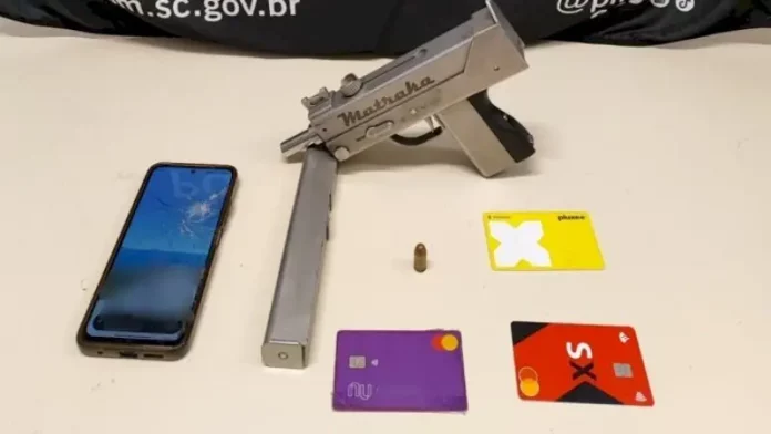 A imagem mostra uma submetralhadora artesanal, um celular e cartões de banco apreendidos durante a operação policial, ilustrando os itens recolhidos pelos agentes.