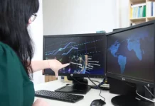 Mulher trabalhando em frente a dois computadores, apontando para uma tela que exibe gráficos, ilustrando análise de dados e atividades no setor de tecnologia.