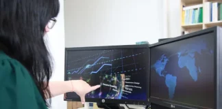 Mulher trabalhando em frente a dois computadores, apontando para uma tela que exibe gráficos, ilustrando análise de dados e atividades no setor de tecnologia.