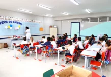 Sala de aula infantil em Florianópolis, com crianças escrevendo em cadernos enquanto a professora escreve no quadro, representando a educação na rede municipal.