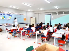 Sala de aula infantil em Florianópolis, com crianças escrevendo em cadernos enquanto a professora escreve no quadro, representando a educação na rede municipal.
