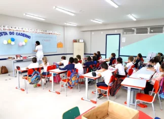 Sala de aula infantil em Florianópolis, com crianças escrevendo em cadernos enquanto a professora escreve no quadro, representando a educação na rede municipal.